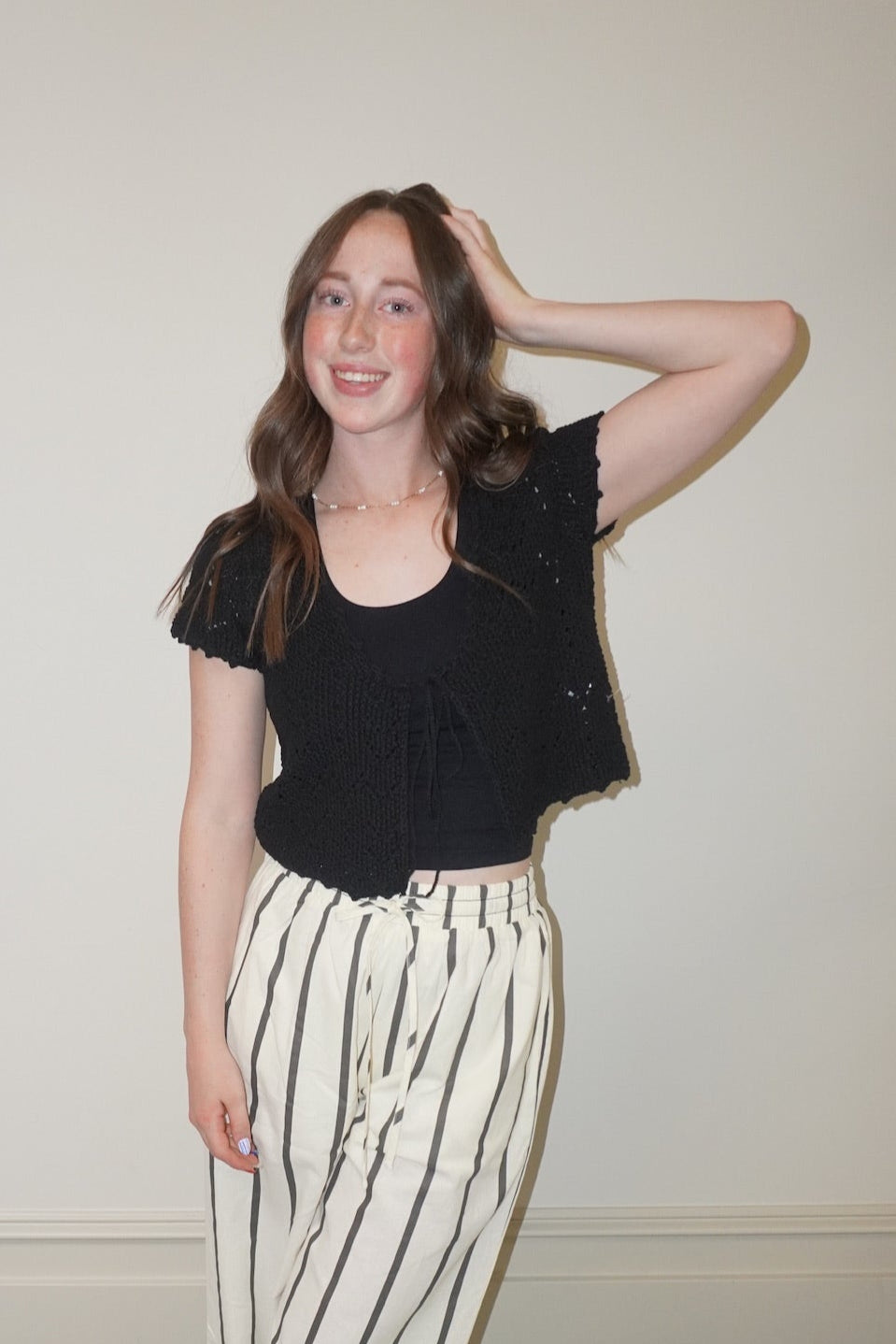 Woman wearing a black knit top and striped pants standing against a white wall.