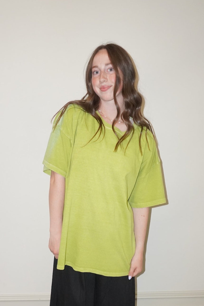 Person wearing a bright green t-shirt and black pants standing against a white wall with a checkered floor.