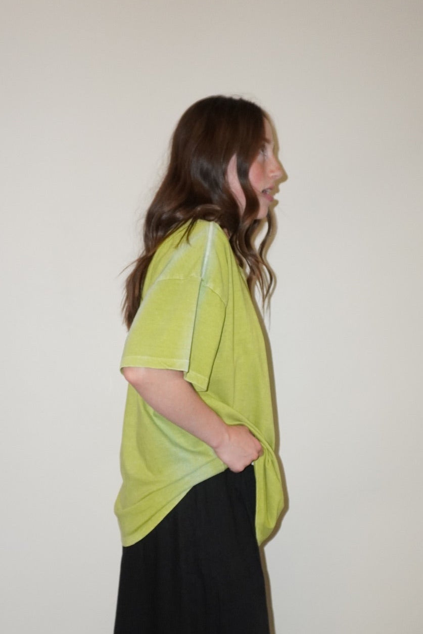 Person wearing a green t-shirt and black pants standing against a white wall with a checkered floor.