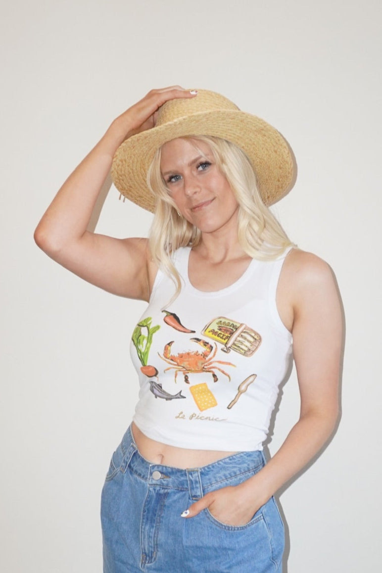 Person wearing a white tank top with graphics, blue denim shorts, and beige hat against a plain wall.