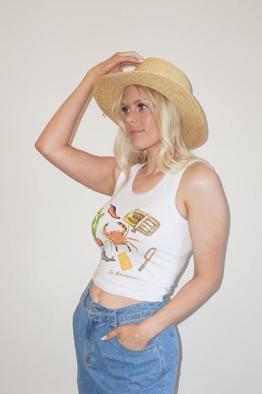 Person wearing a white tank top with graphics, blue shorts, and a straw hat against a plain wall.