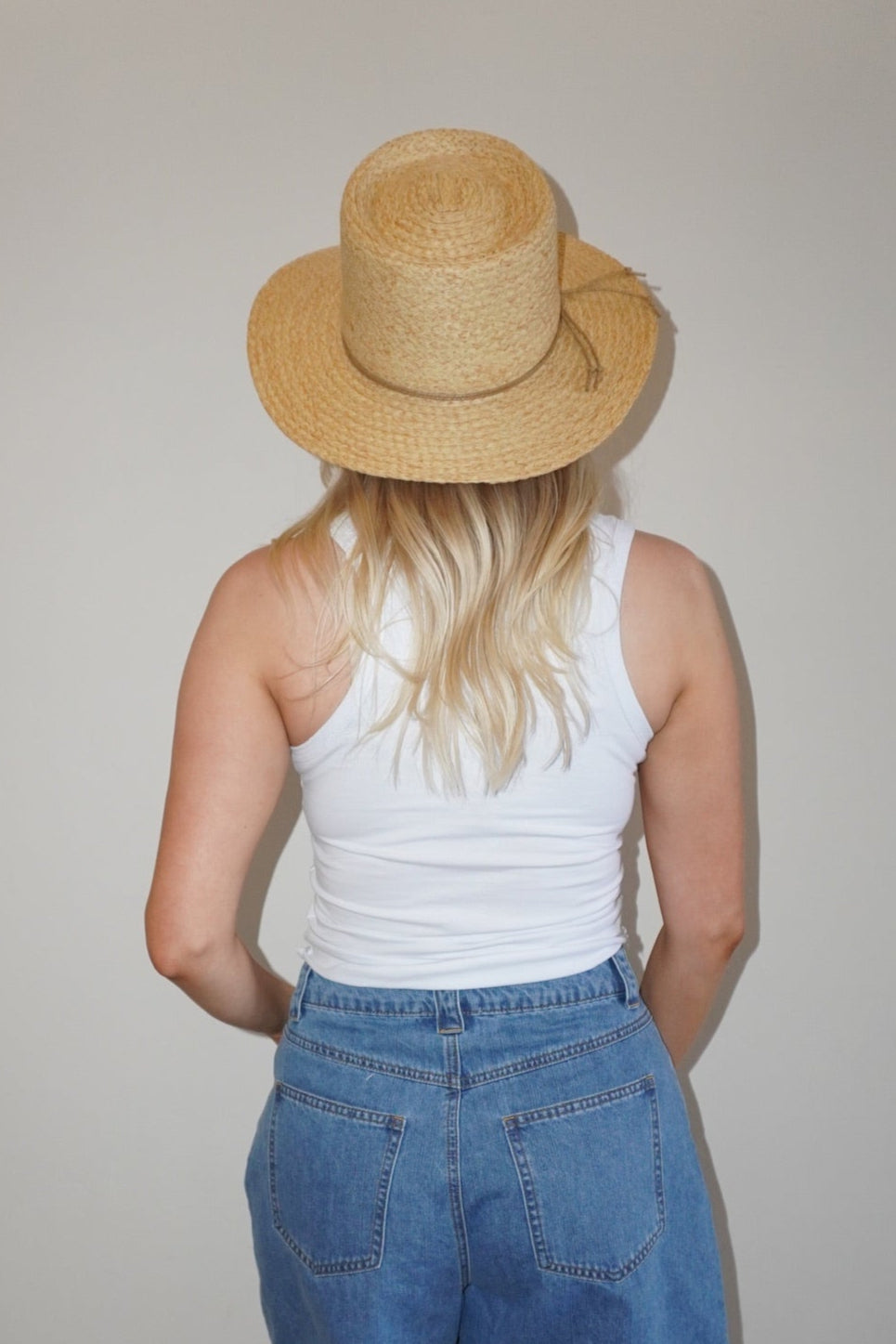 Person wearing a straw hat, white tank top, and blue denim shorts standing against a plain wall.