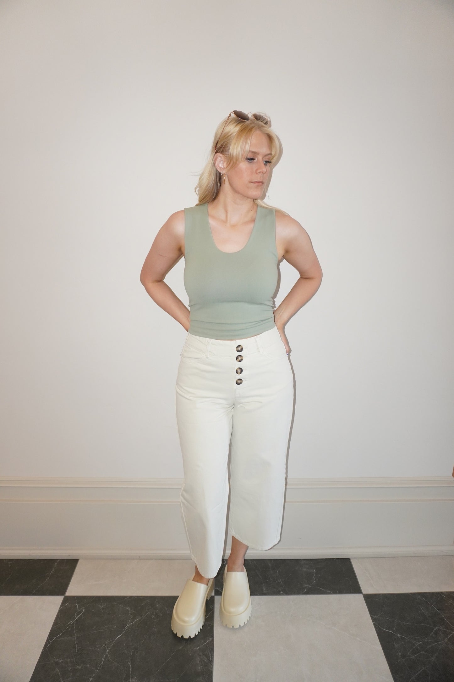 Woman wearing a green tank top and white pants standing against a plain wall.