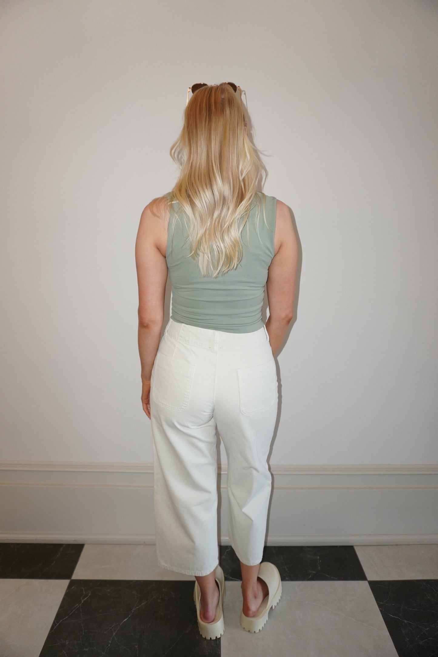 Person wearing a green tank top and white pants standing against a plain wall.