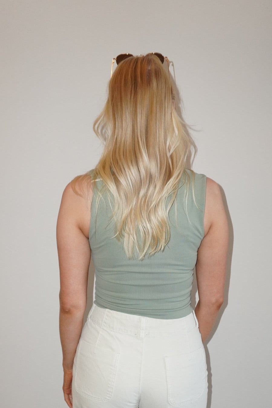 Person wearing a green tank top and white pants standing against a plain wall.