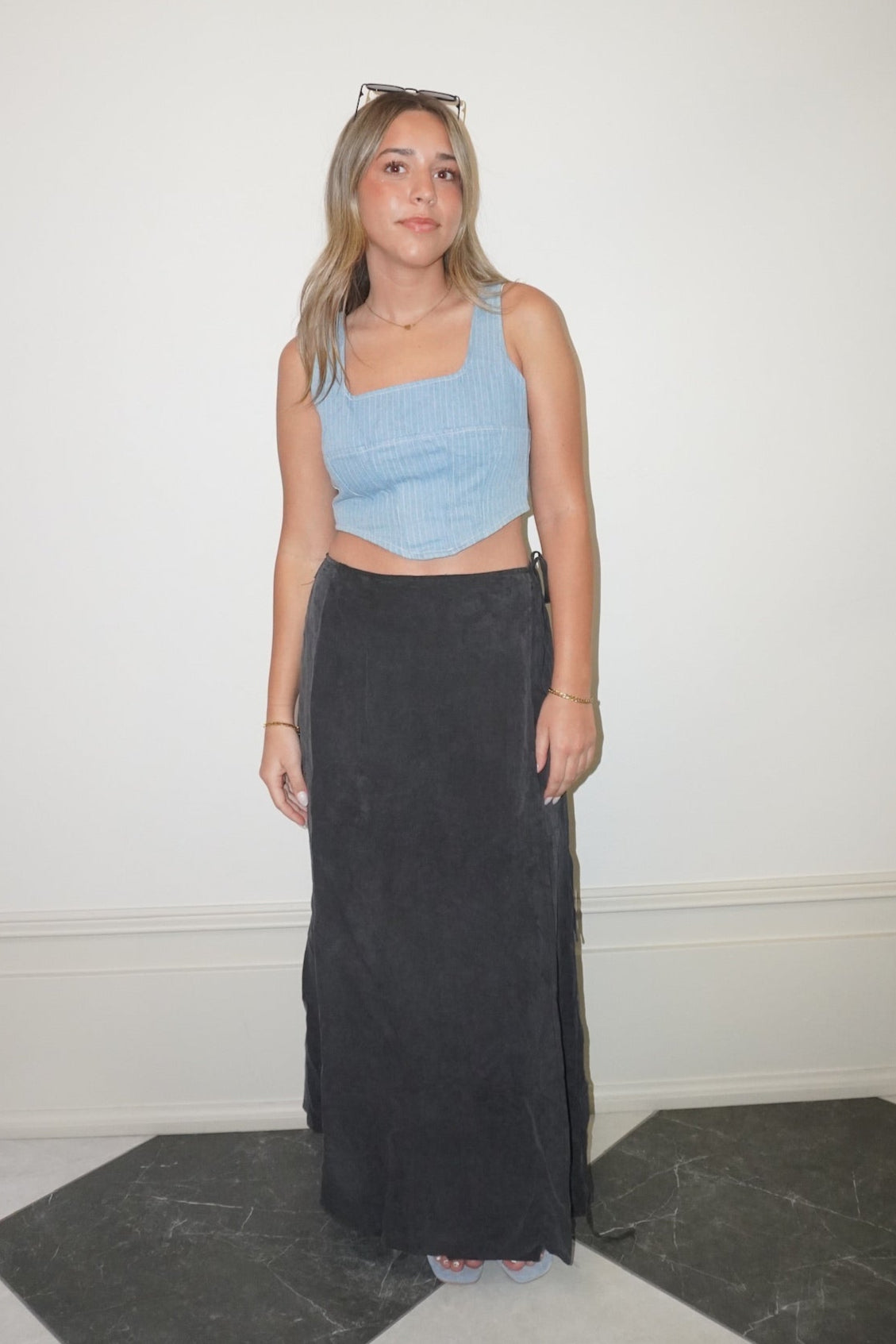 Woman wearing a blue crop top and black skirt standing in a room with a white wall and checkered floor.