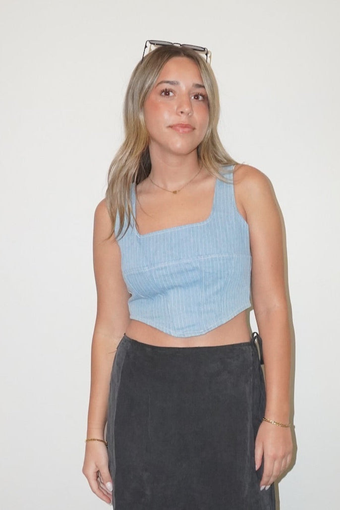 Woman wearing a blue crop top and black skirt standing in a room with a white wall and checkered floor.