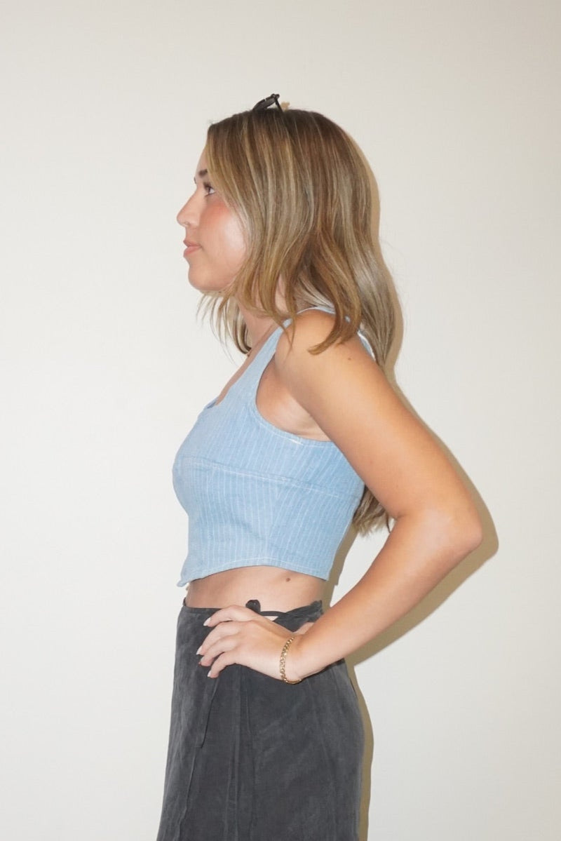 Woman wearing a light blue crop top and dark gray wide-leg pants standing against a white wall.