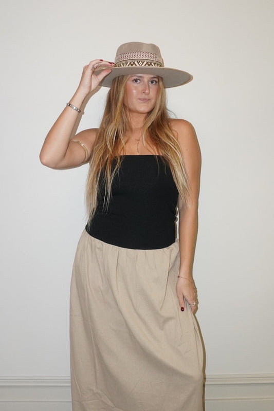 Person wearing a black top and beige skirt holding a hat against a white wall.