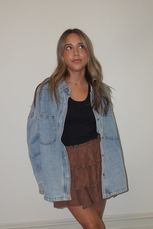 Woman wearing a denim jacket, black top, and brown skirt standing against a white wall.