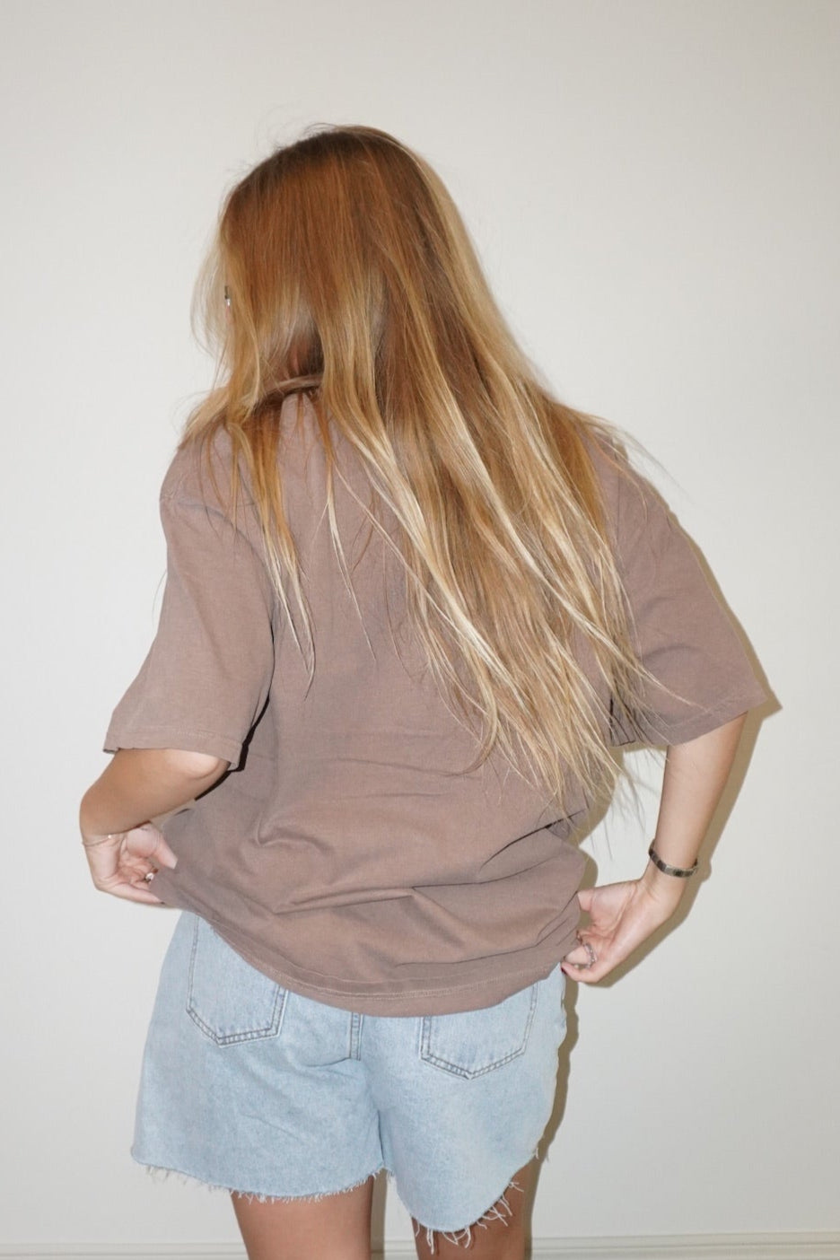 Person wearing a brown oversized shirt and light blue denim shorts standing against a white wall.