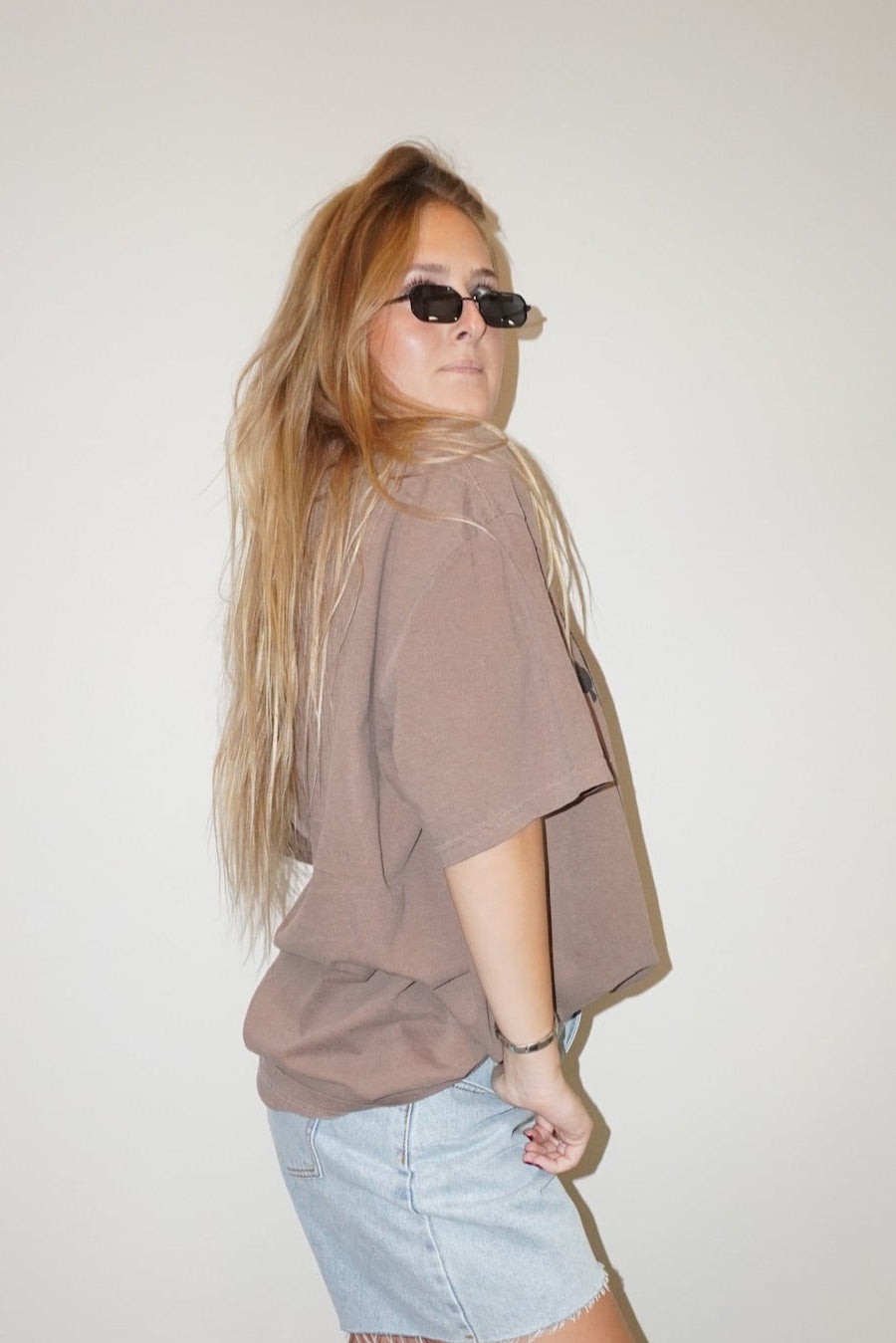 Woman wearing a brown oversized shirt and light blue shorts against a plain wall.