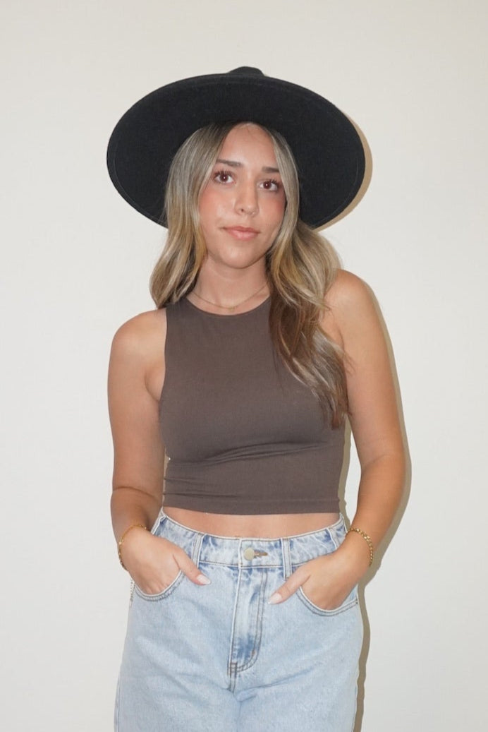 Person wearing a brown tank top, light blue denim skirt, black hat, and black sandals on a white wall background.