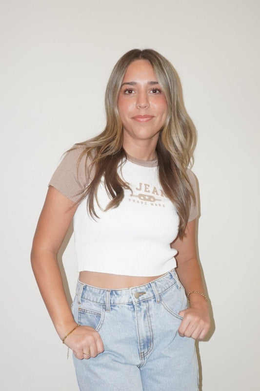 Woman wearing a white crop top and denim shorts standing against a plain wall.