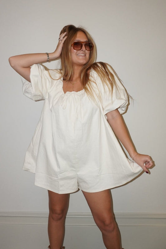 Woman in a white outfit with brown boots posing against a plain wall.