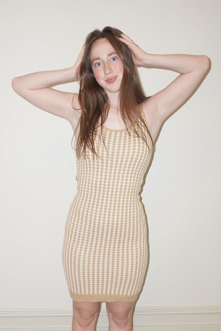 Woman wearing a beige crochet like dress standing against a white wall.