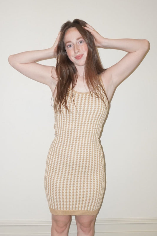 Woman wearing a beige crochet like dress standing against a white wall.