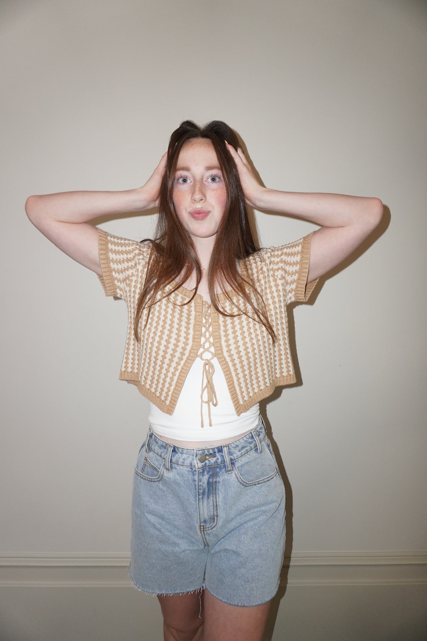 Person wearing a beige knitted cardigan over a white top and denim shorts against a plain background