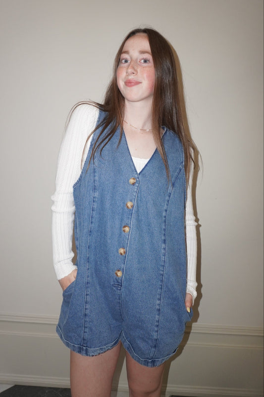 Person wearing a denim romper over a white shirt against a plain wall.