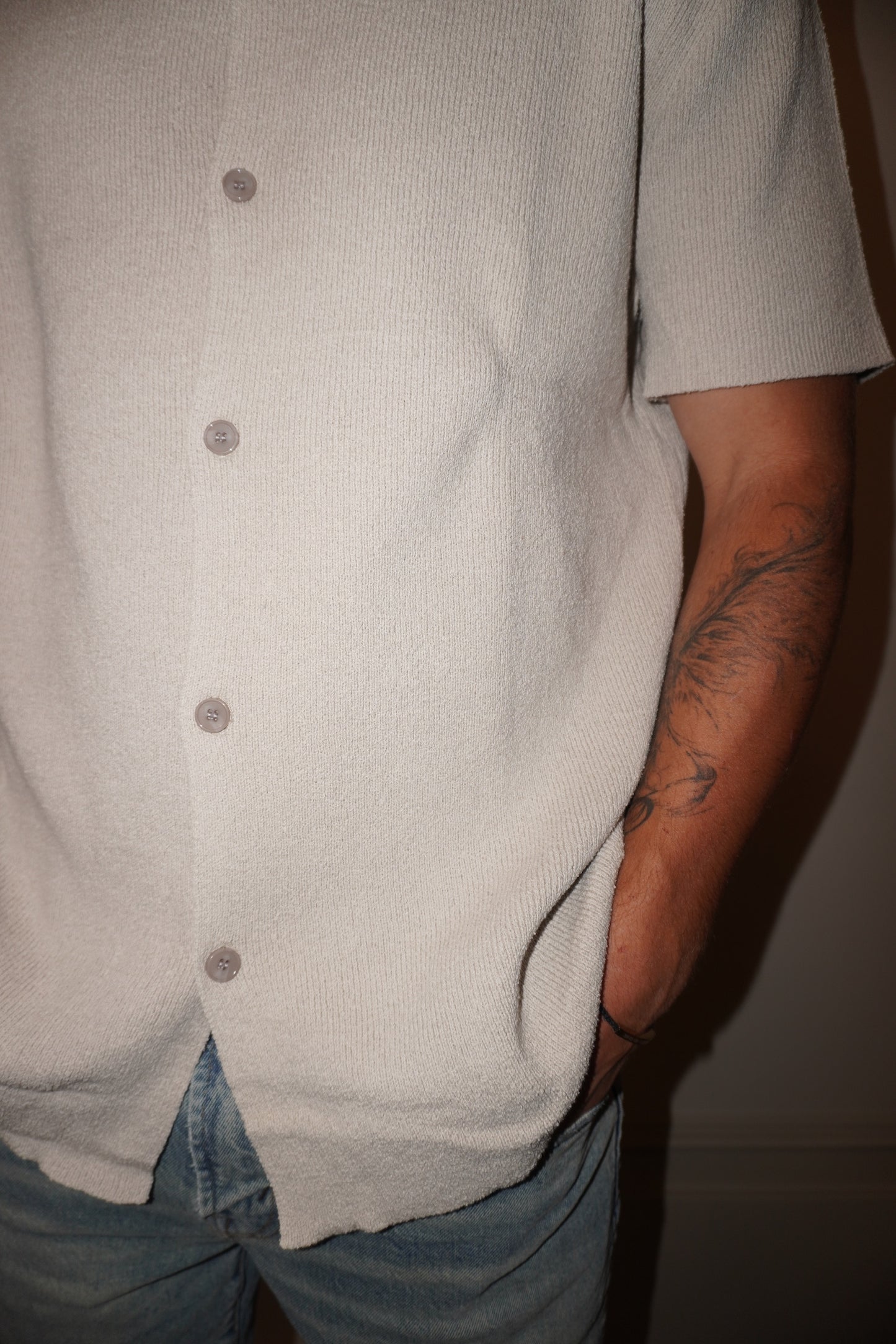 Person wearing a light gray button-up shirt and blue jeans with a tattoo on the arm.