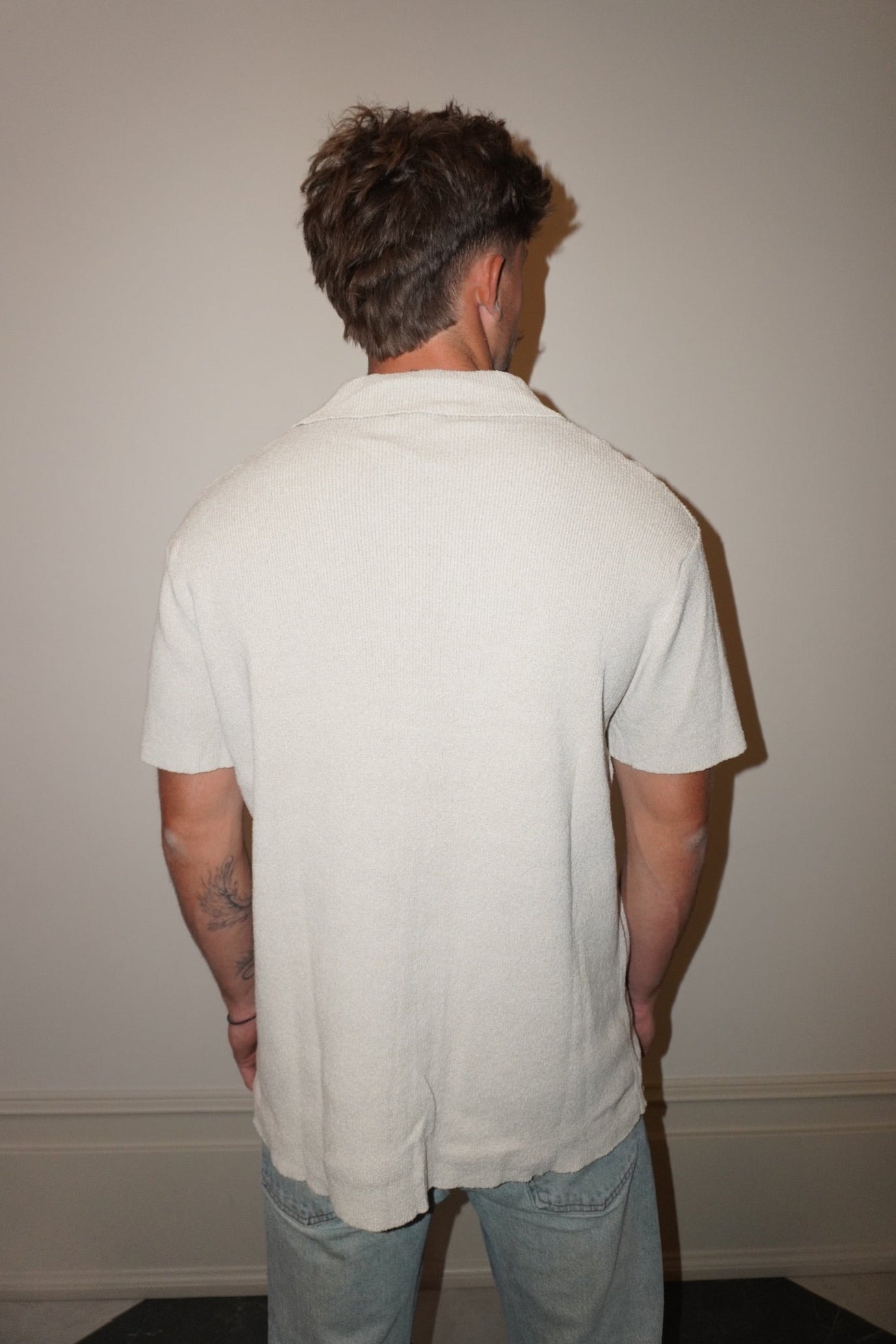 Man wearing a light-colored shirt and jeans standing against a plain wall.