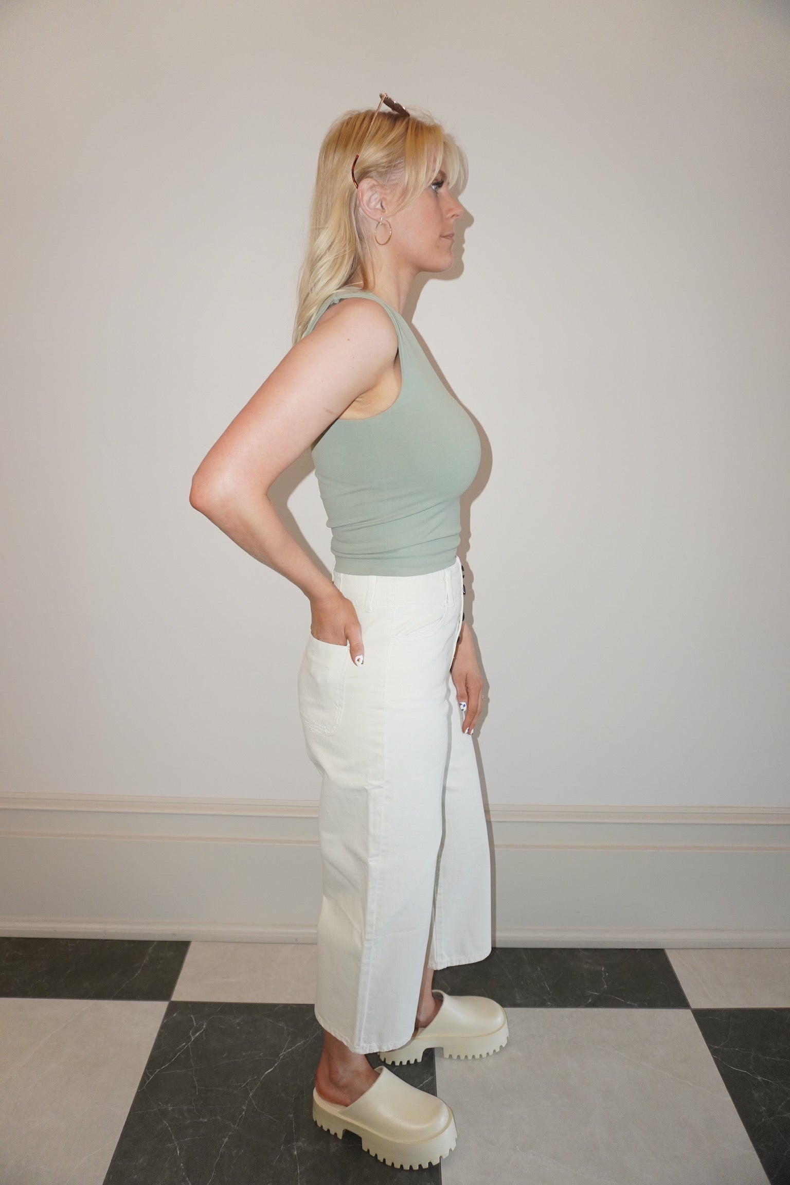 Woman wearing a green top and white pants standing against a plain wall.
