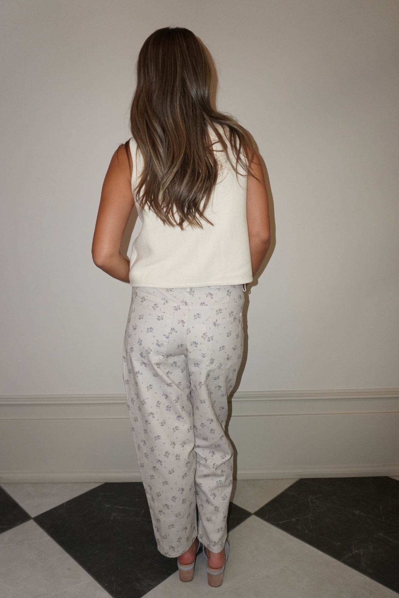 Person wearing a white sleeveless top and light gray pants with a subtle pattern, standing in a room with a checkered floor.