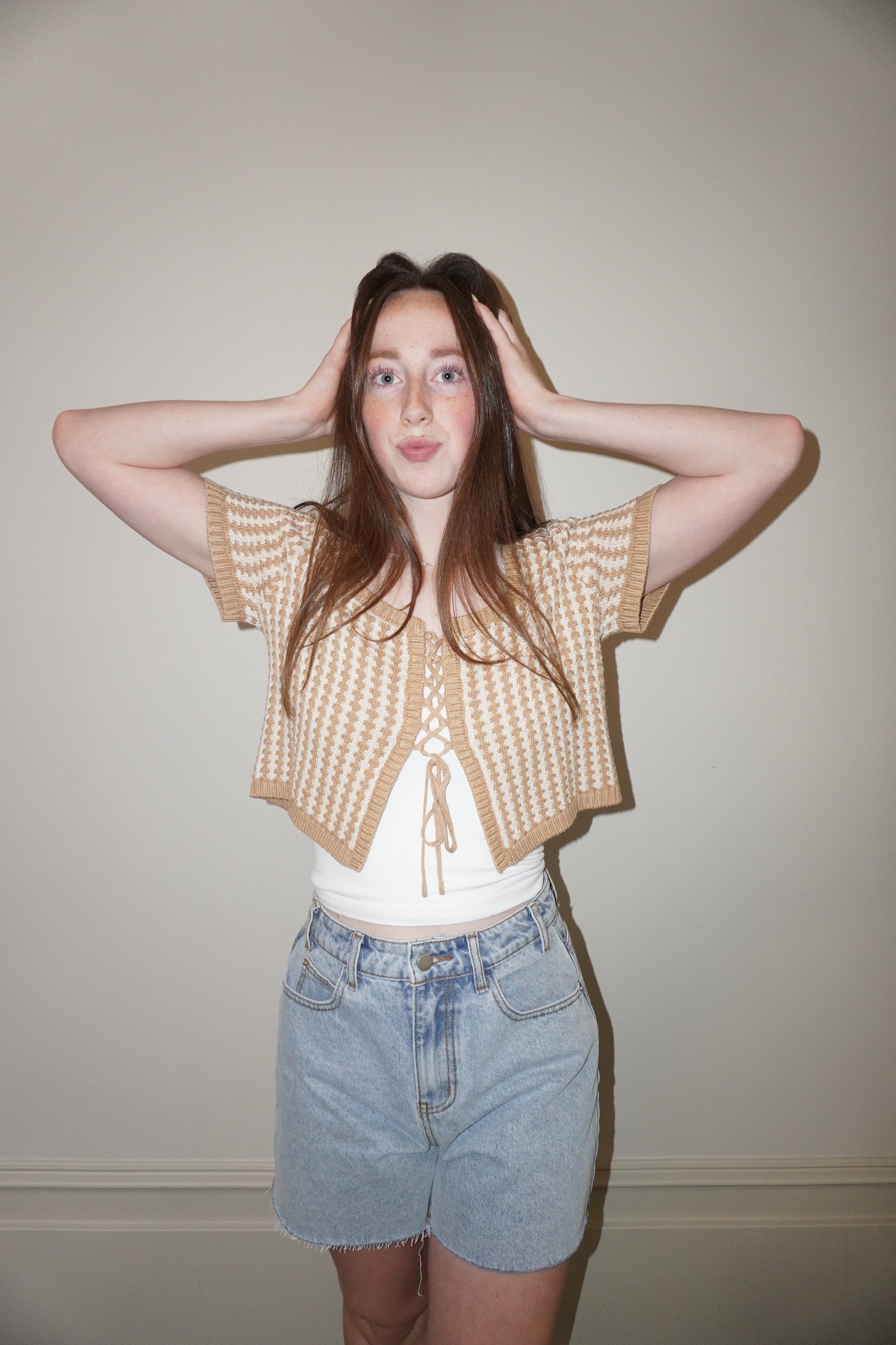 Person wearing a beige knitted cardigan over a white top and denim shorts against a plain background