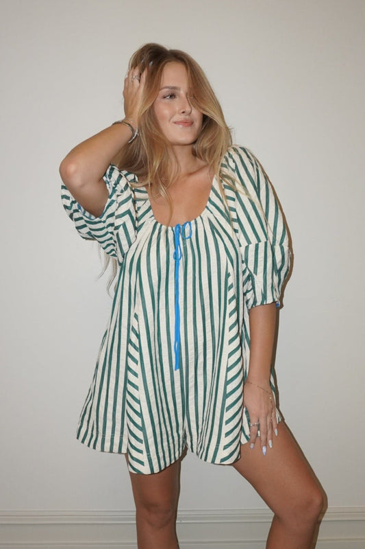 Woman wearing a green and white striped romper with a blue zipper, standing against a plain wall.